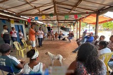 Grupo de pessoas reunidas em círculo sob uma estrutura coberta, com cadeiras plásticas ao redor, algumas pessoas em pé falando ao grupo e um cachorro no centro do espaço. Bandeirolas coloridas estão penduradas no teto.