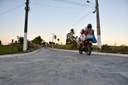 Pessoa em motocicleta trafegando por estrada pavimentada com blocos de concreto, em área rural, com postes de energia, cercas, vegetação e árvores ao longo da via.