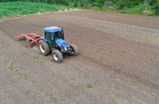 Trator agrícola azul puxando implemento de preparo do solo em área de plantio, com terreno arado e vegetação ao fundo.